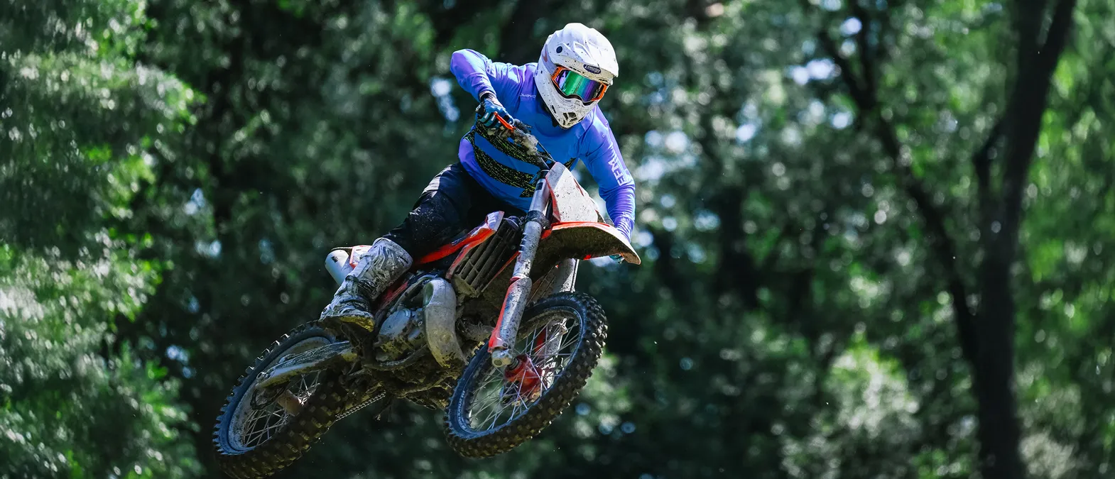 A motorcyclist in a blue long-sleeved racing suit and white helmet jumps on a motorcycle through a forest.