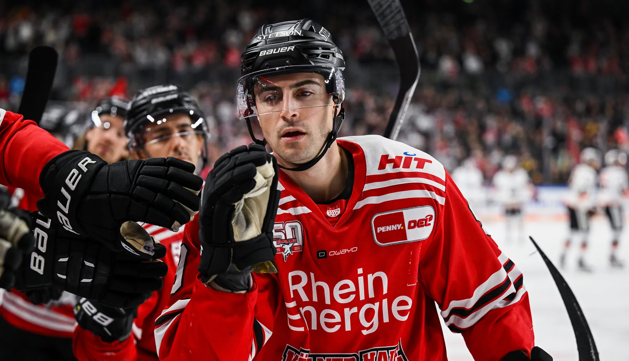 A hockey player in a red jersey with white stripes and gloves, reacting during the game.