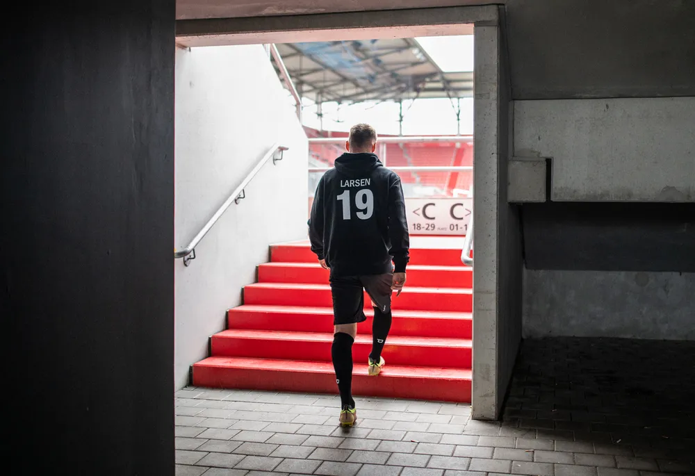 A soccer player in a black hoodie with the number 19 and shorts is walking up the red steps of a stadium.