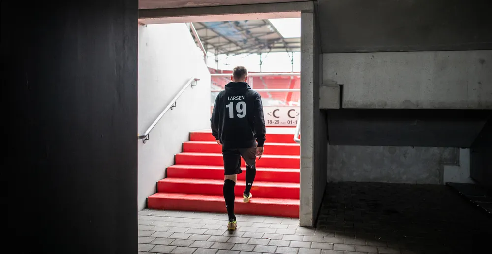 An athlete in a black hoodie with the number 19 and black shorts walking up a red staircase in a stadium.