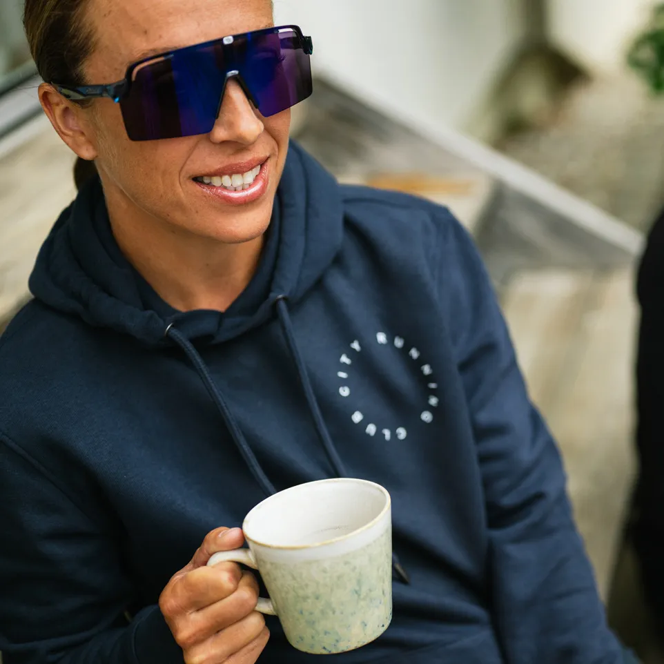 A person wearing a dark blue hoodie with a logo is smiling at the camera while holding a cup.