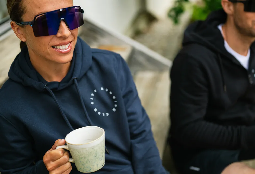 A person wearing a blue hoodie with a logo holds a cup while sitting on a wooden bench.