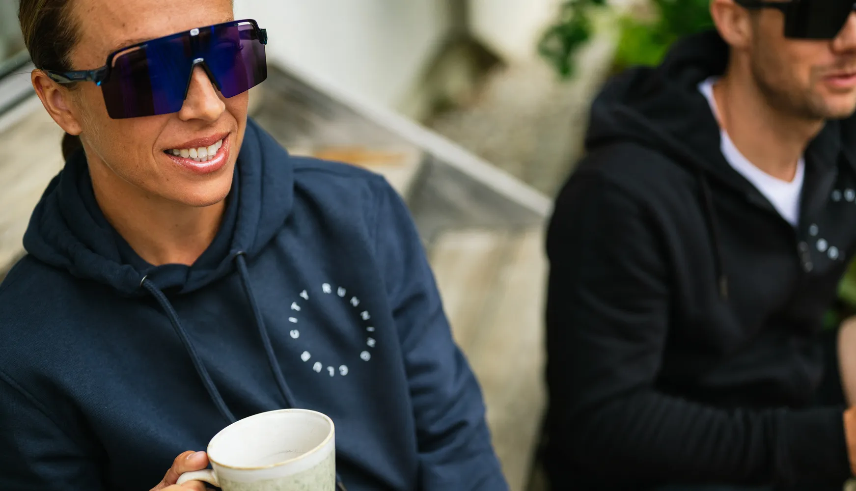 A person in a blue hoodie is holding a cup and wearing large sunglasses, while another person in the background is also wearing a hoodie.