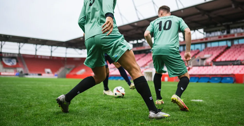 Two soccer players in light green jerseys and black socks training on a grass field in a stadium.