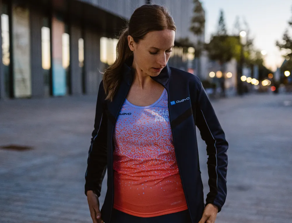 A woman in athletic clothing, consisting of a lightweight jacket and a colorful top, stands in an urban setting, looking thoughtfully at her outfit.