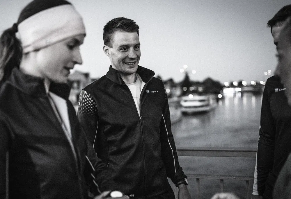 A group of athletes wearing dark sports jackets stands by the water's edge during dusk.