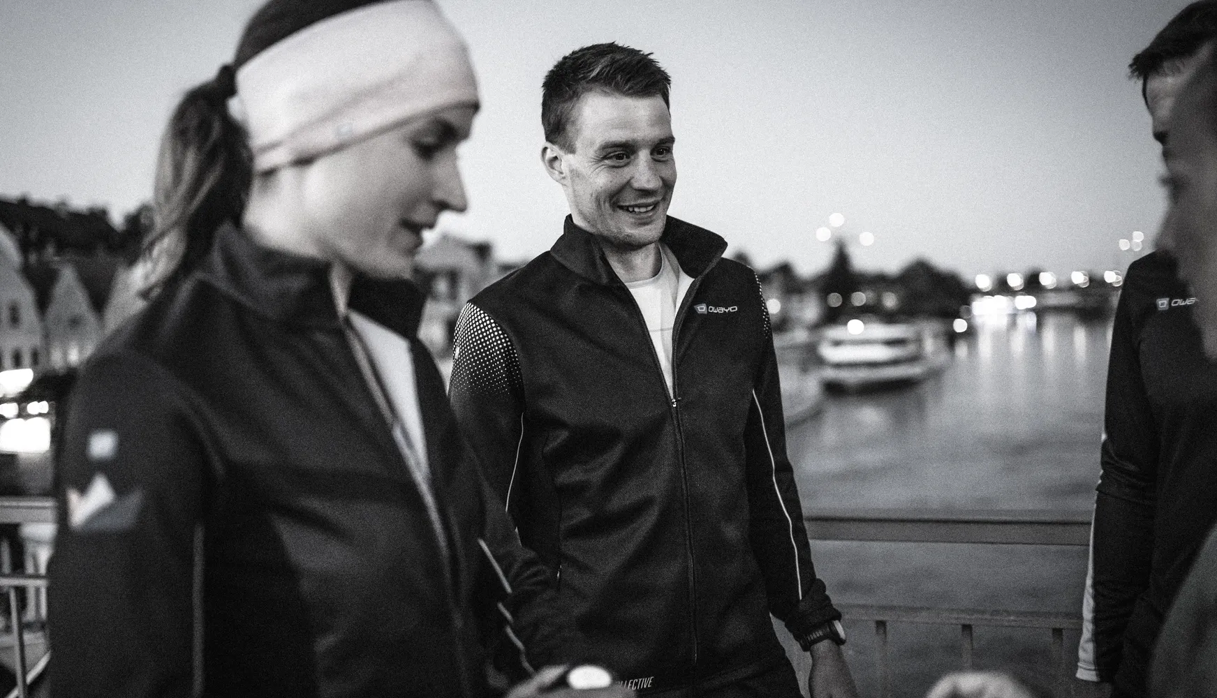 Two people in sporty jackets stand by the waterfront, one wearing a headband and looking thoughtfully, while the other smiles.