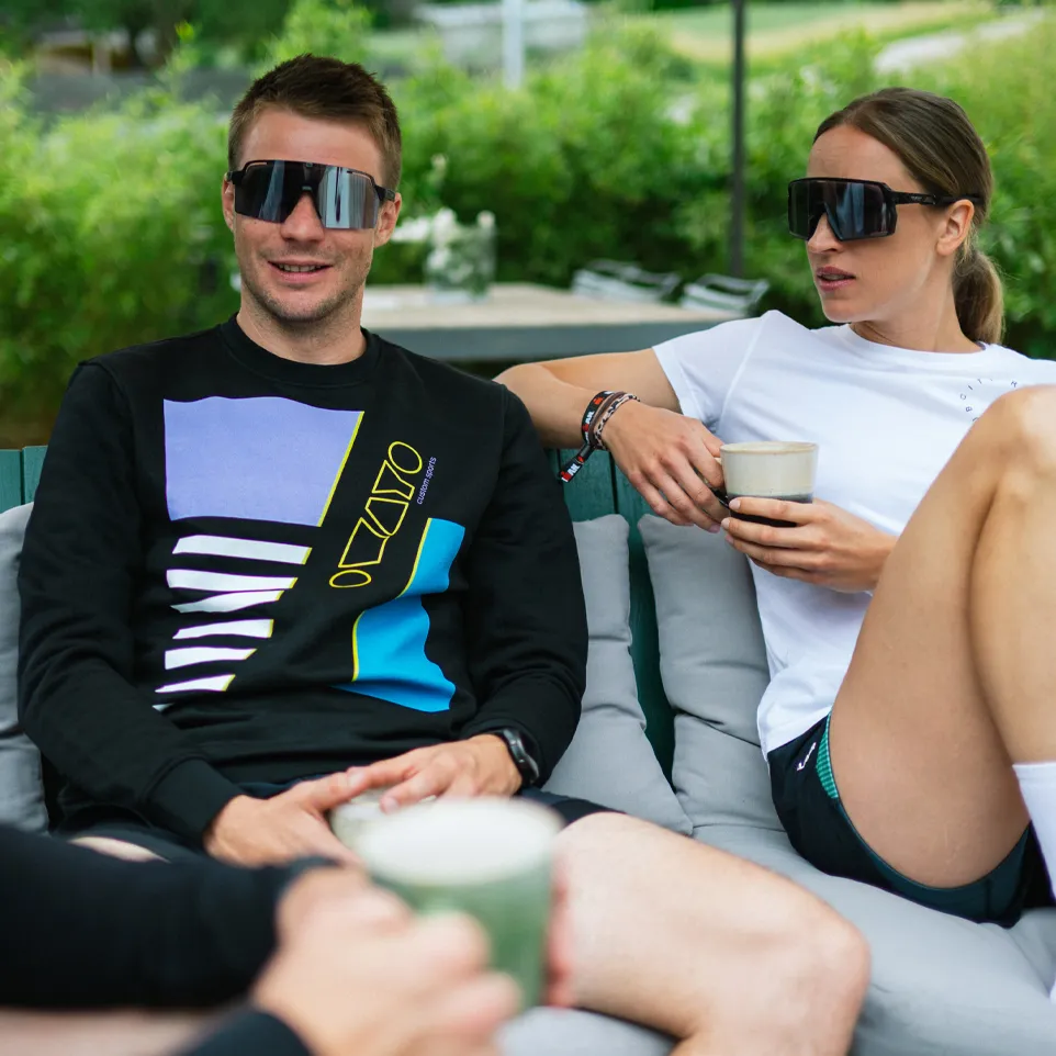 Two people sit on an outdoor couch, the man wearing a black sweatshirt with a colorful design and the woman in a white t-shirt, both holding drinks.