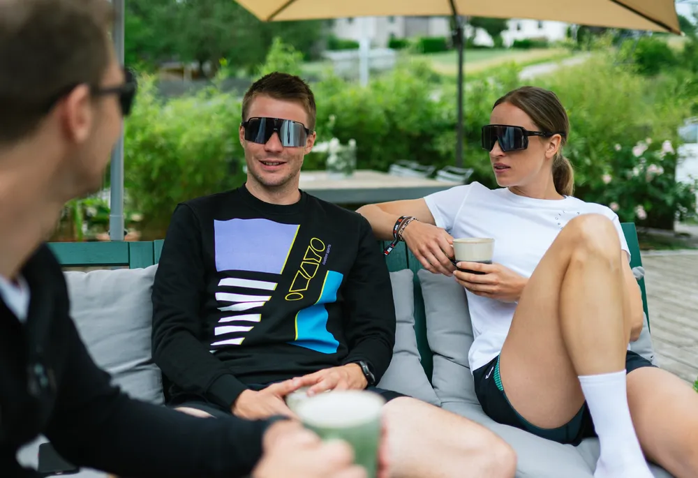 Three people on a couch, two wearing casual sportswear and sunglasses, while they relax and chat, holding drinks.