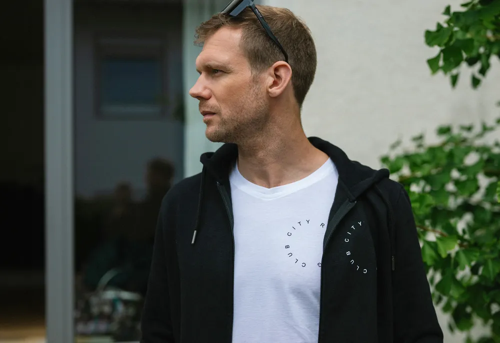 A man stands sideways wearing a black hoodie and a white t-shirt featuring a circular print.