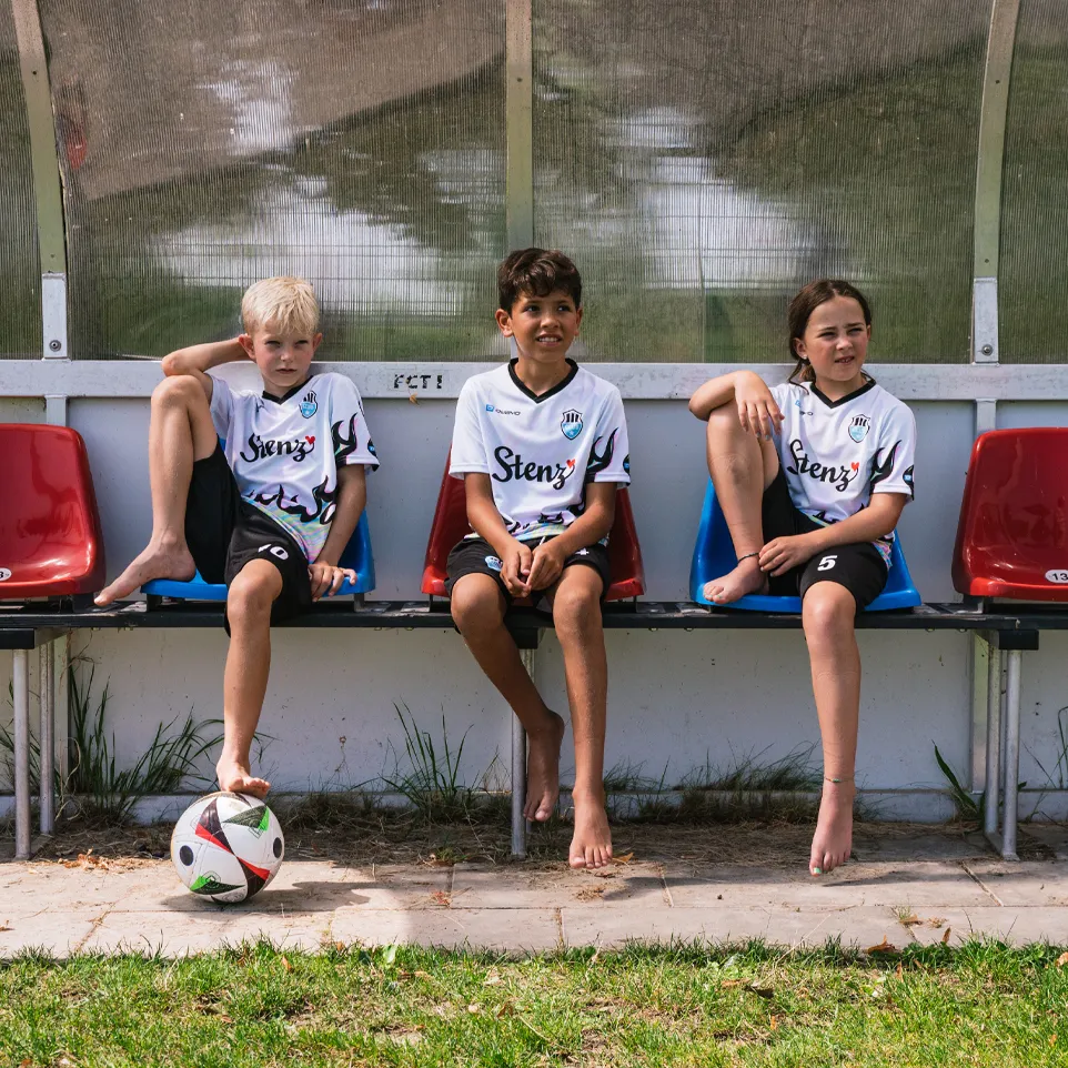 Three children in soccer jerseys and shorts sit on a bench by the field, with a soccer ball in front of them.