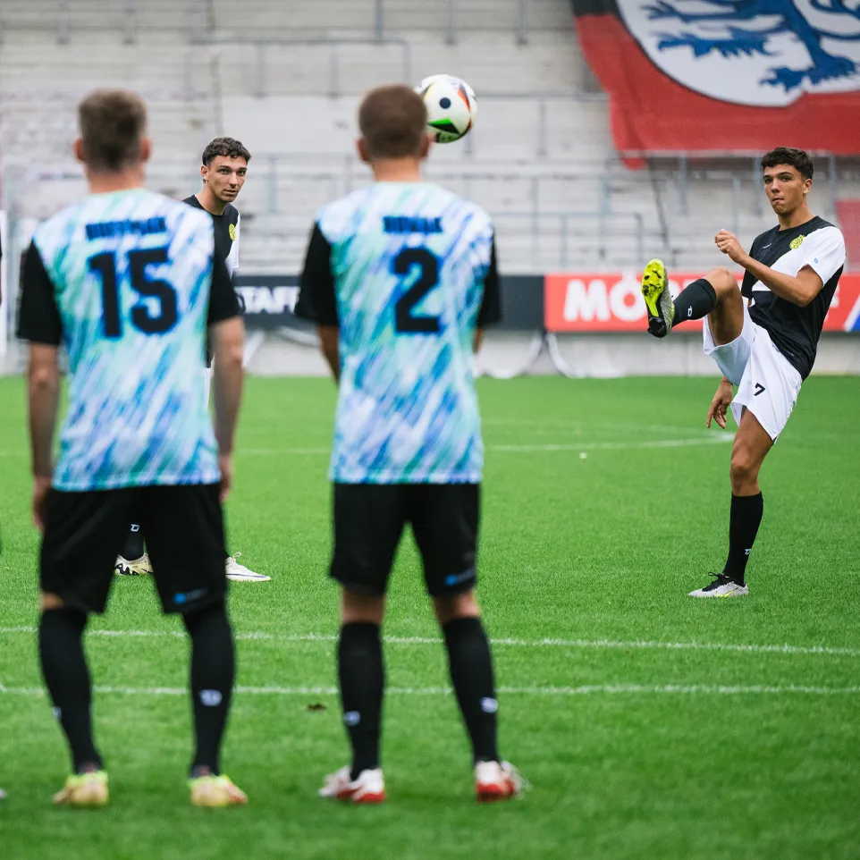 Three football players in colorful jerseys stand on the field watching another player juggling a ball.