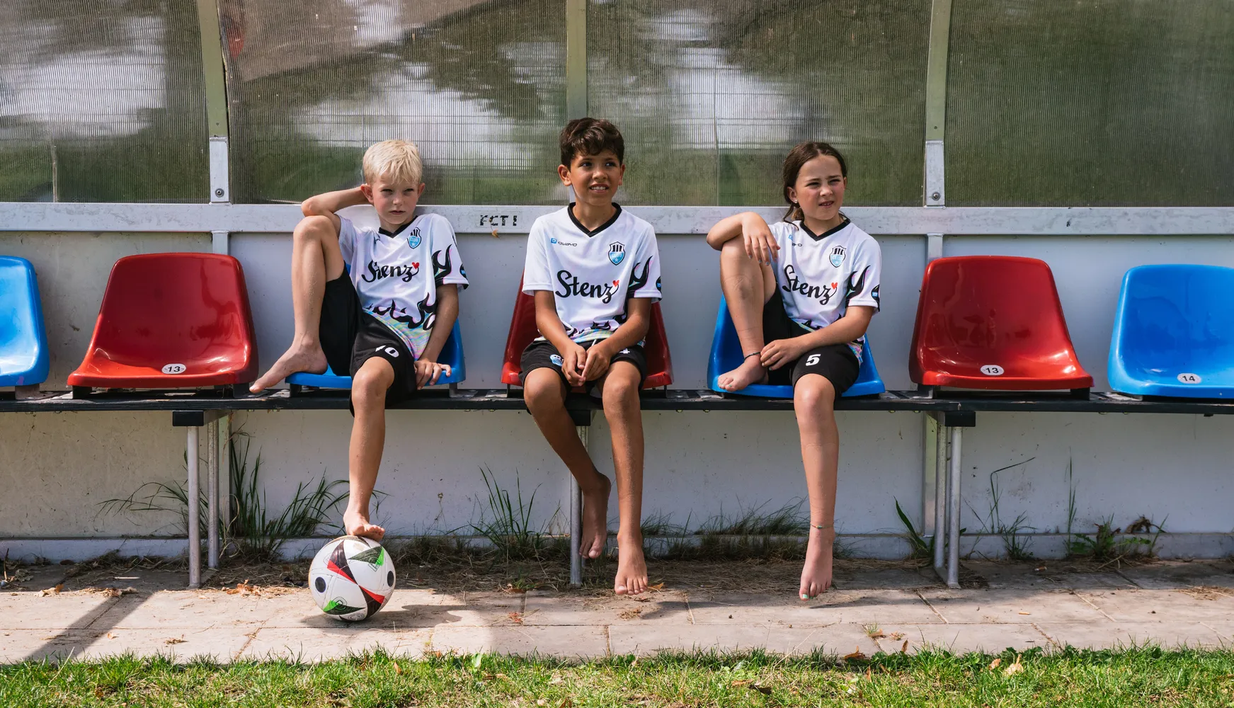 Three children in customized soccer jerseys are sitting on a bench with a soccer ball on the ground.