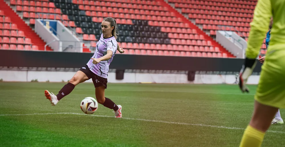 A female player in a purple and white soccer jersey kicks the ball on a stadium field, with a goalkeeper in yellow gear in the background.