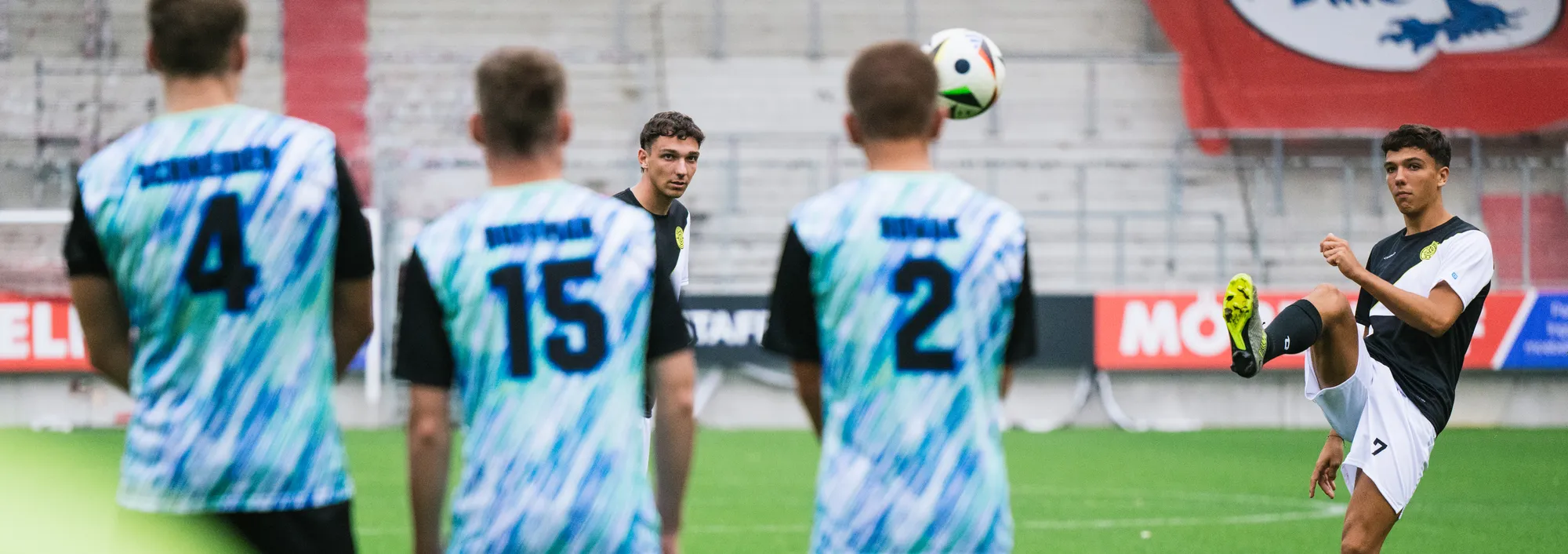 A group of soccer players in colorful jerseys stands on the field while one of them lifts a ball with their foot.