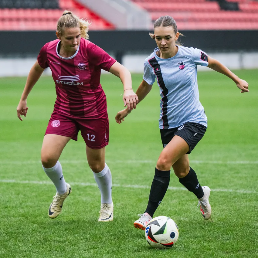 Two women in football jerseys are playing on a grass field, attempting to control the ball.