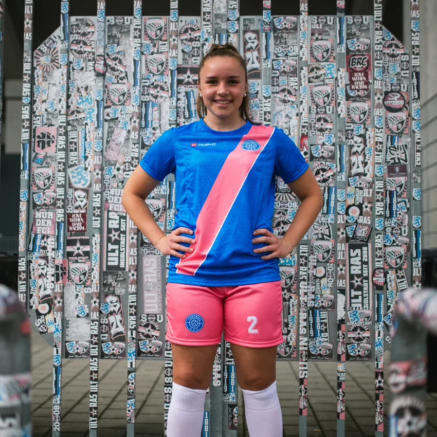 A girl smiles in front of a wall covered in stickers, wearing a blue soccer jersey with a pink stripe and pink shorts.