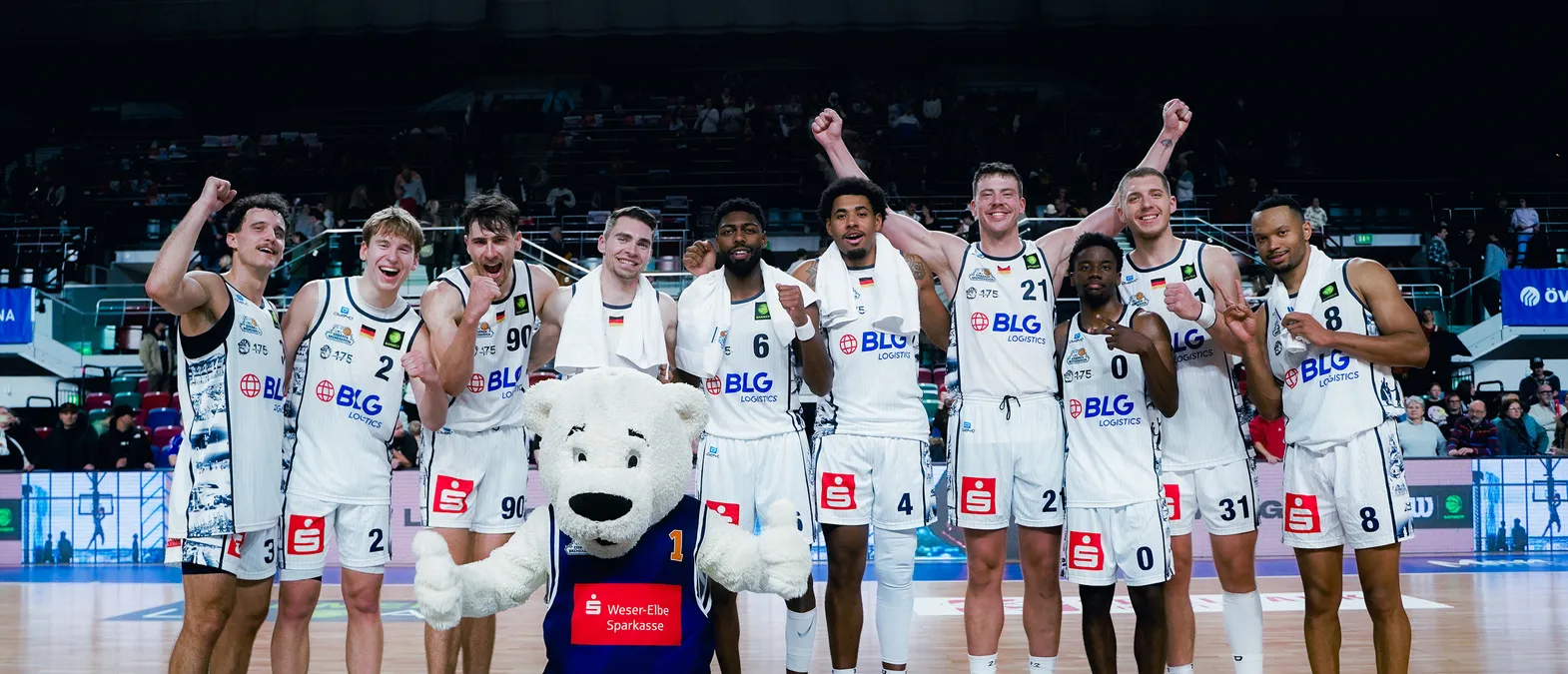 Basketball players in white jerseys with the BLG Logistics logo celebrate on the court, surrounded by a crowd, with a polar bear mascot in the foreground.