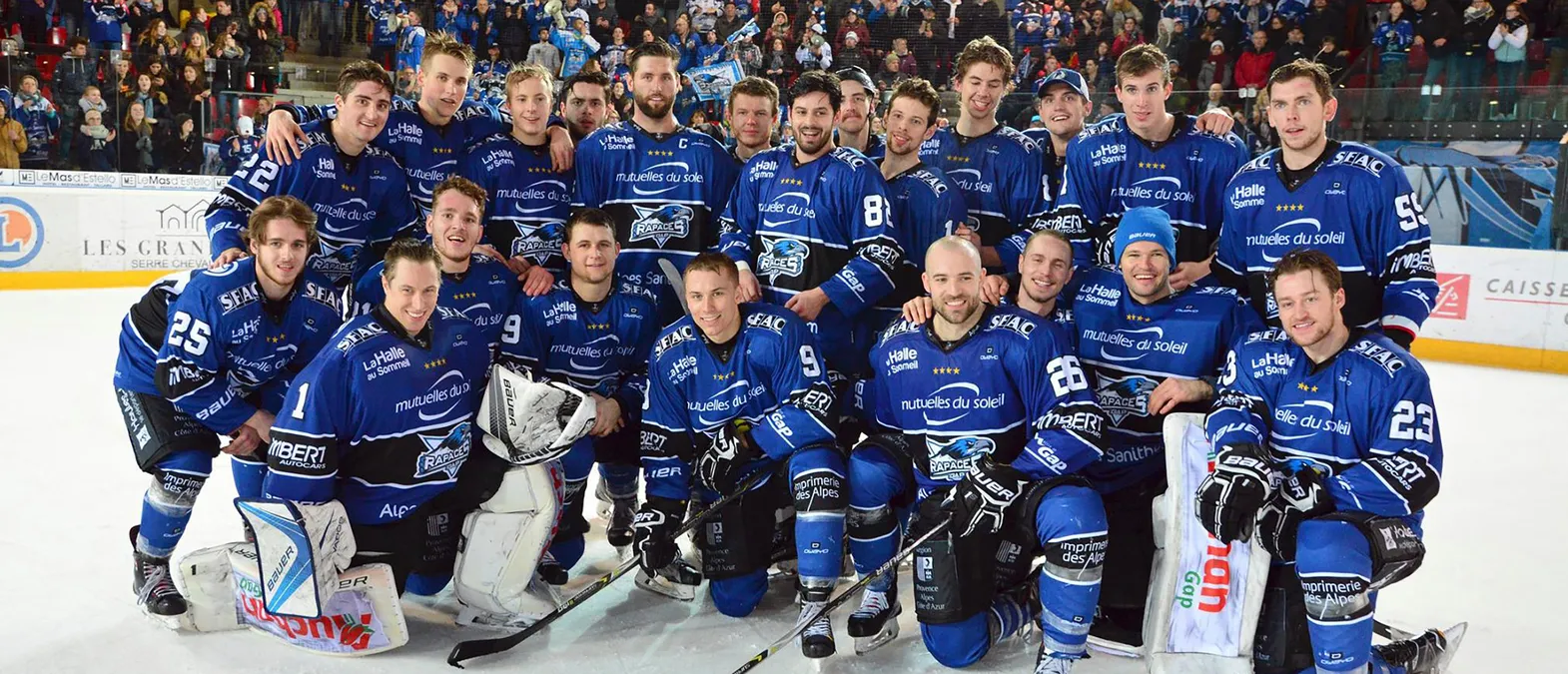 A hockey team in blue jerseys with the Rapaces de Gap logo poses on the ice in front of a crowd.