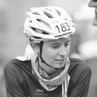 A female cyclist wearing a black cycling jersey with the 'IWC Schaffhausen' logo, a white helmet with the number 183, and a scarf around her neck.