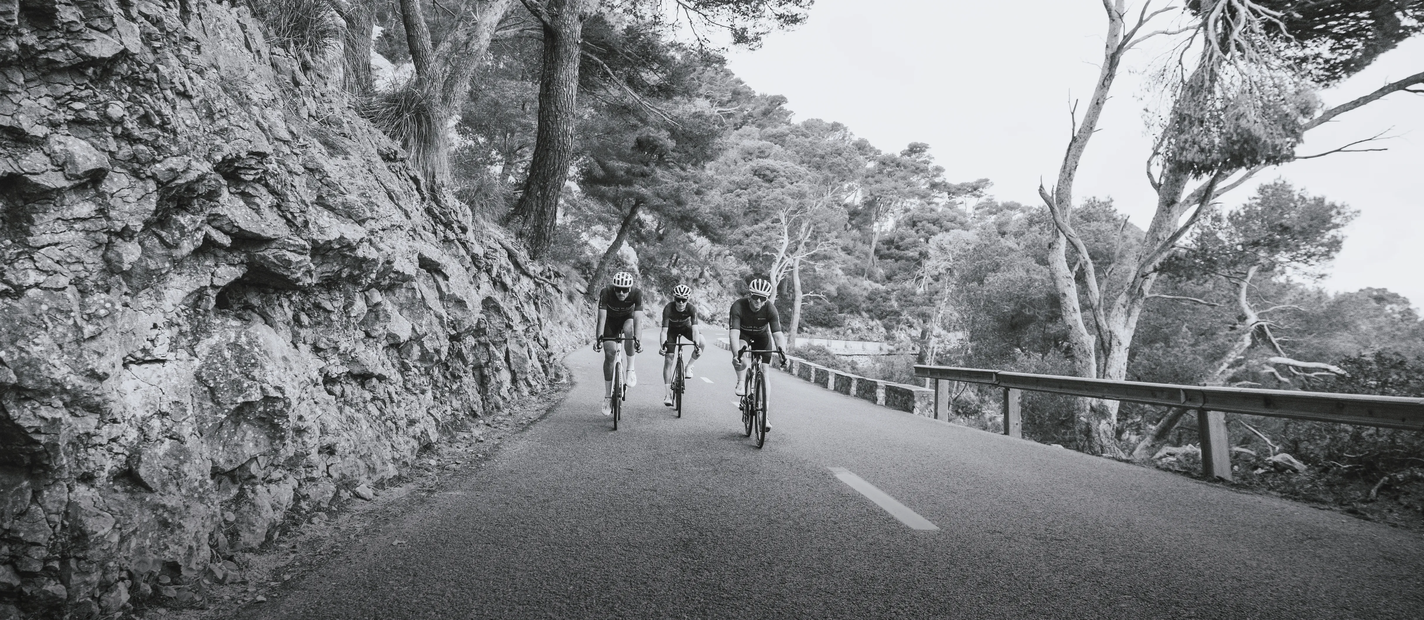 Three cyclists in sportswear ride on a road alongside a rocky cliff.