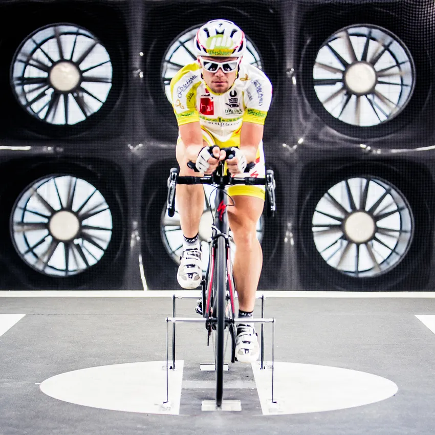 A cyclist in a yellow and white jersey rides on a stationary bike, surrounded by large fans.