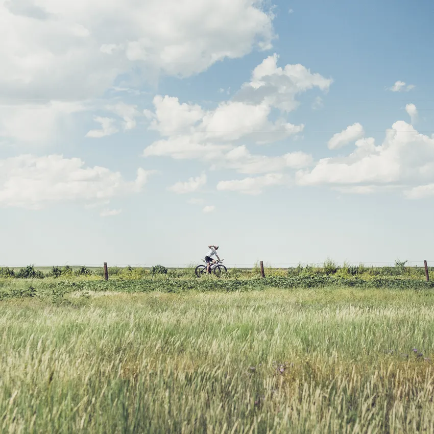 A person rides a bicycle through a green landscape under a blue sky with some clouds.