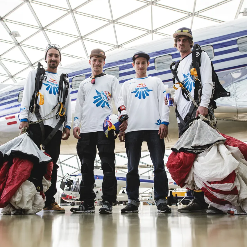 Four people in sporty clothing stand in a modern hangar, ready for a skydiving jump.