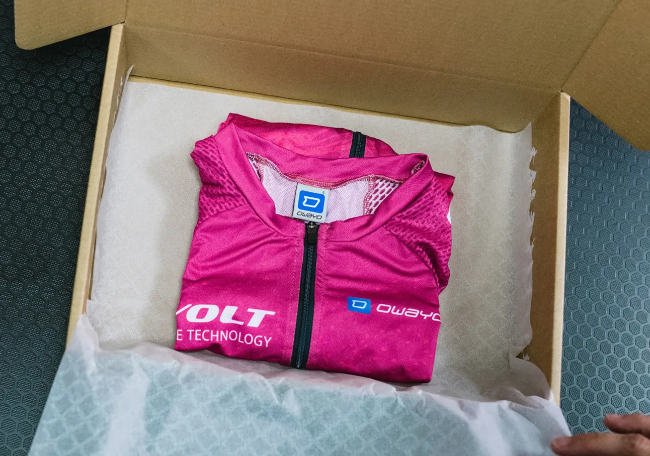 A folded pink sleeveless sports jersey with the owayo logo is placed in a cardboard box on white paper.